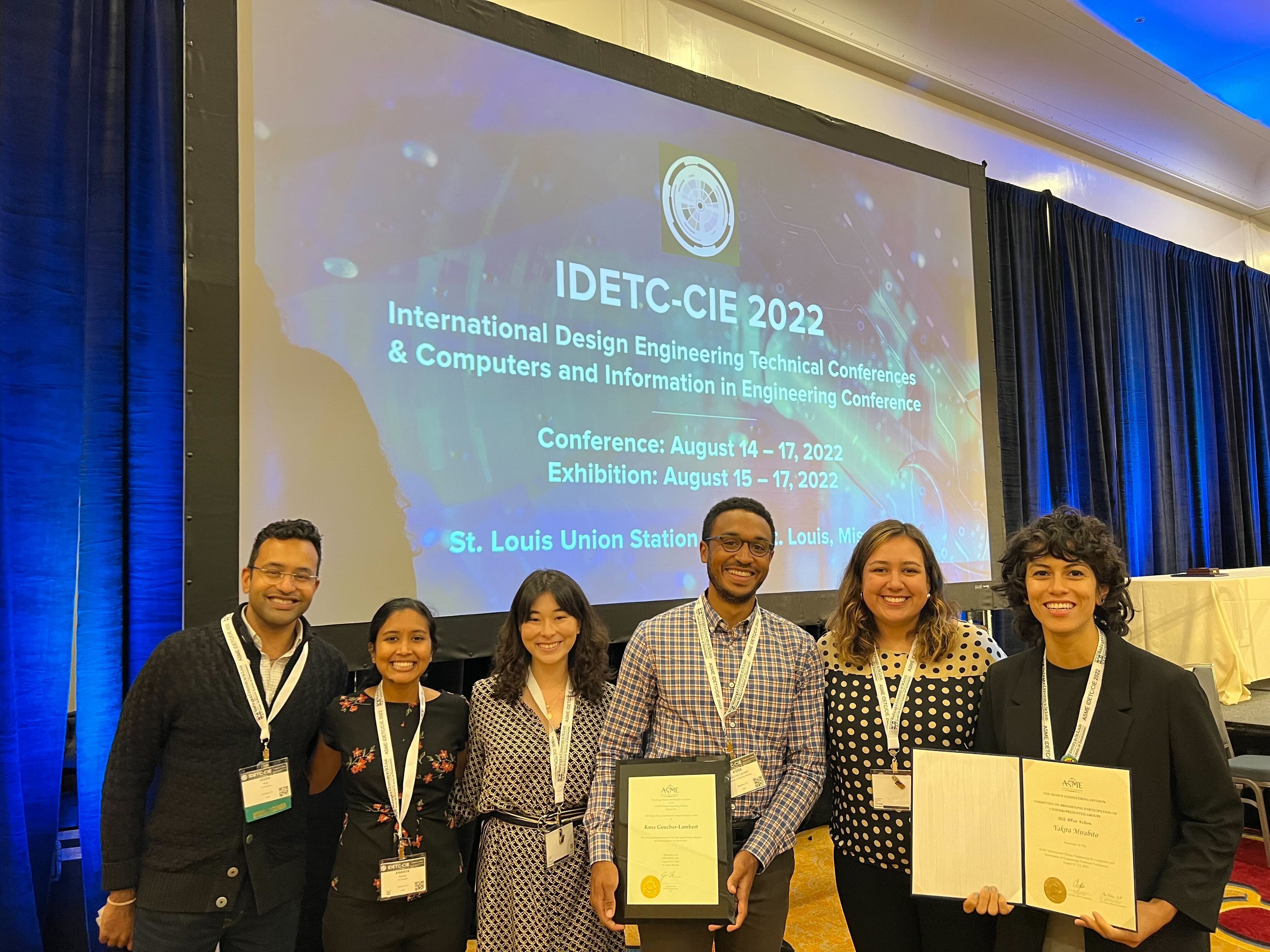 Vivek, Ananya, Elisa, Kosa, holding award, Nicole, and Yakira, holding certificate, in front of screen with IDETC conference title
